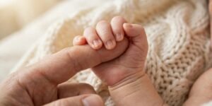 Newborn baby's tiny hand holding a parent's finger on a cream knitted blanket.